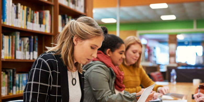 Tre personer arbetar vid ett bord i ett bibliotek. Foto.