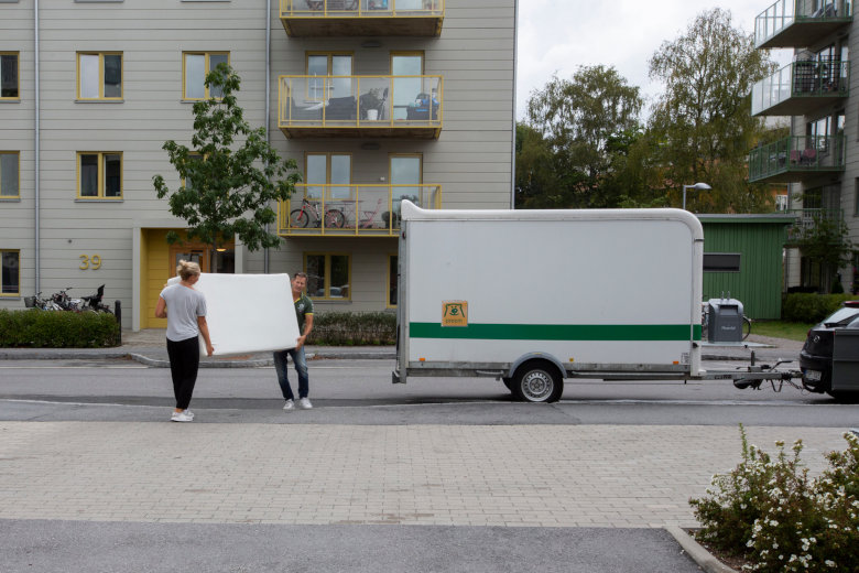 Två personer bär en stor vit skiva framför ett hyreshus, med en flyttbil parkerad på gatan bredvid. Foto.