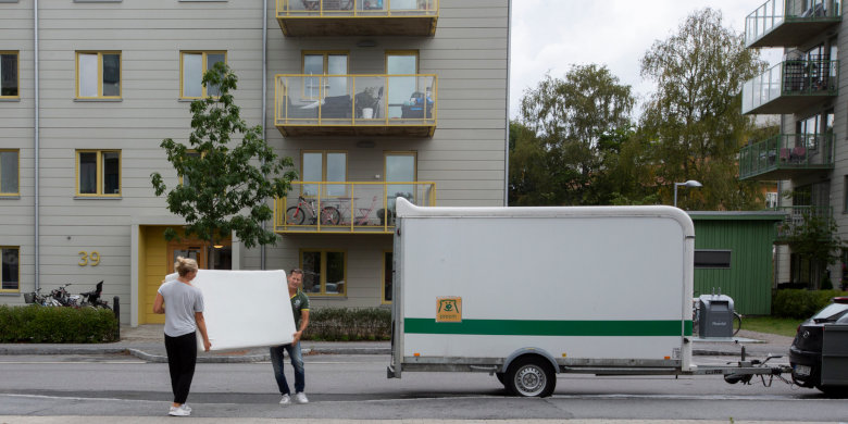 Två personer bär en stor vit skiva framför ett hyreshus, med en flyttbil parkerad på gatan bredvid. Foto. 
